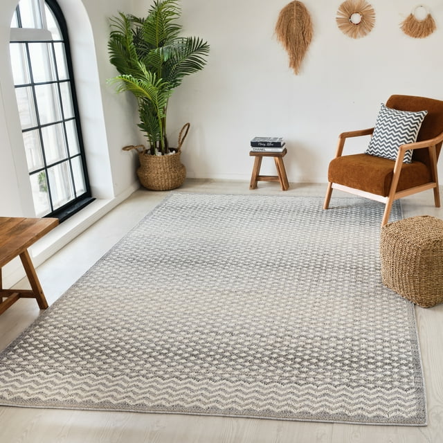 Grey beige Moroccan geometric patterned indoor area rug on light wood floor in a bright living room with brown armchair, woven basket planter, small wooden stool with books, and natural fiber wall decor.