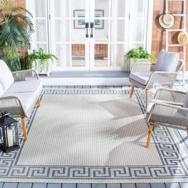 SAFAVIEH Bermuda Isidora machine washable indoor/outdoor area rug featuring a blue and white geometric Greek key border design in a modern sunroom with wicker furniture and plants.