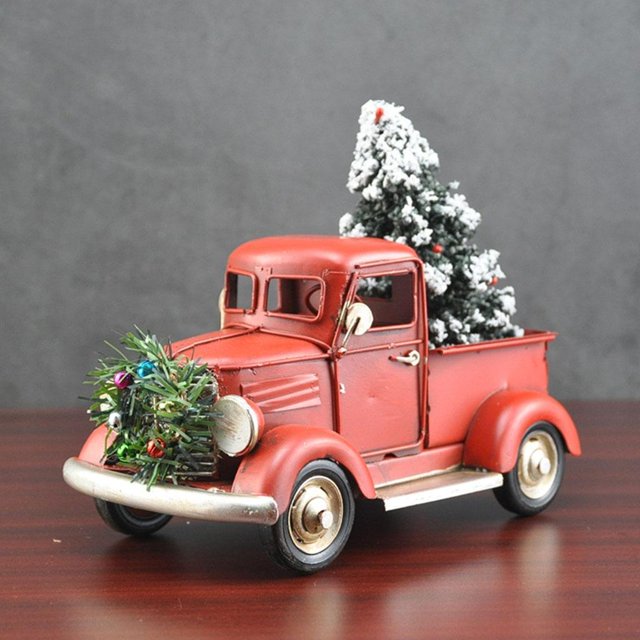 decoración de navidad colcomx camión rojo 21x11x16cm adorno navideño de hierro, vista frontal centrada de camioneta roja clásica con caja trasera que lleva un árbol de Navidad con nieve artificial, pintura roja mate, parachoques y repisas plateadas, ruedas con detalle dorado y corona verde con pequeñas esferas en la parrilla; fondo gris neutro, ideal para repisas, puertas o colgar en el árbol.