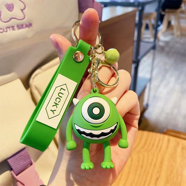 llavero de juguete de mickey y minnie de disney, mike wazowski, james p. llavero colgante de dibujos animados, llaveros de puntada, regalo de cumpleaños para niña Llaverito frontal verde Mike Wazowski de goma espuma con un ojo grande y sonrisa dentada blanca y negra; brazos y piernas cortos, anilla y cadena metálica dorada y correa de silicona verde marcada 'LUCKY', sostenido sobre mesa de madera en interior. También suele usarse como adorno para mochilas o bolsas pequeñas.