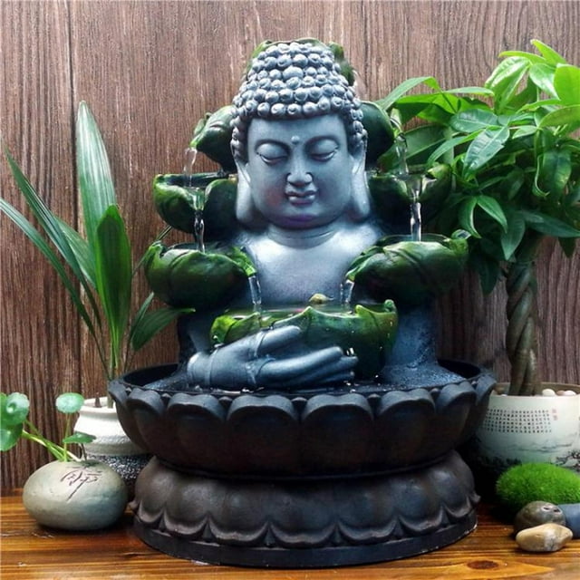 Elodie Sitting Buddha Tabletop Outdoor Garden LED Lights Desktop Waterfall with a brown base. The Buddha statue is crafted from resin, featuring cascading water flowing through green leaf-like tiers, illuminated by soft LED lights. The fountain is placed on a wooden tabletop surrounded by potted plants and decorative stones, creating a tranquil and meditative ambiance. Ideal for home, office, or outdoor patio decor.