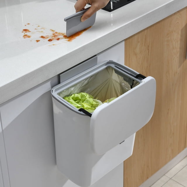 cubo de basura grande creativo montado en la pared para cocina con tapa cubo de basura doméstico con tapa deslizante cubo de basura para baño para inodoro tipo colgante, vista en 3/4 frontal de un cubo colgante crema blanco con tapa abatible abierta y bolsa interior visible, montado bajo la encimera junto al fregadero para desechar restos de comida; también suele usarse para organizar residuos de baño o papelería ligera.
