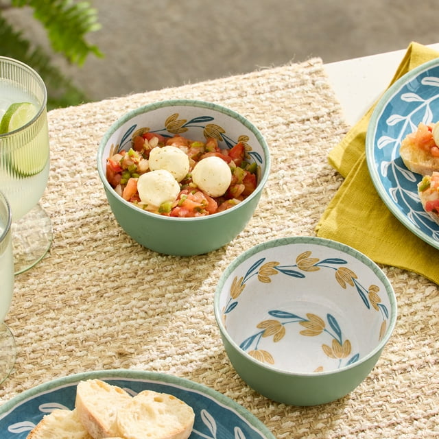 Better Homes & Gardens Bamboo Melamine Flower Dip Bowl 4.65 Inch 2Pc set with two green bowls featuring yellow and blue floral patterns inside, one filled with tomato salsa and mozzarella balls, placed on a woven placemat with plates of bread and glasses of lime water, suitable for serving sauces, snacks, or condiments.