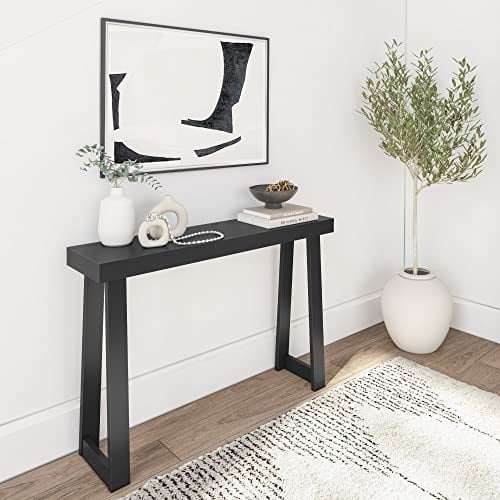 Plank+Beam Solid Wood Console Table, 46.25-inch narrow entryway table in black, shown in a modern living room setting. The table features a sleek rectangular top supported by sturdy angled legs. It is styled with decorative items, including a vase, books, and a tray, and placed against a white wall with framed artwork above. A potted plant and a textured rug complete the scene.