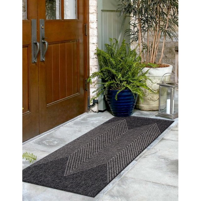 ECARPET Raven contemporary runner rug with chevron design in dark tones, displayed on an entrance stone floor beside wooden double doors and decorative plants.