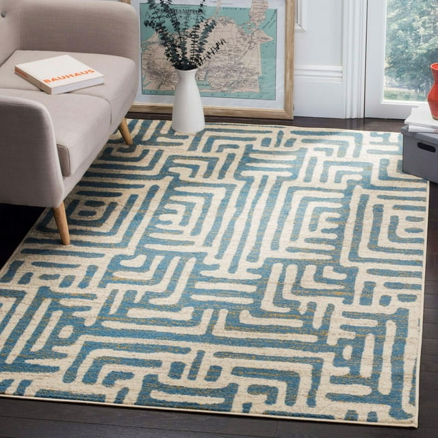 SAFAVIEH Amsterdam Elvan geometric area rug in blue and beige tones placed on dark hardwood floor beneath a modern light gray sofa, accented by decorative elements including a white vase with eucalyptus branches and framed vintage map.