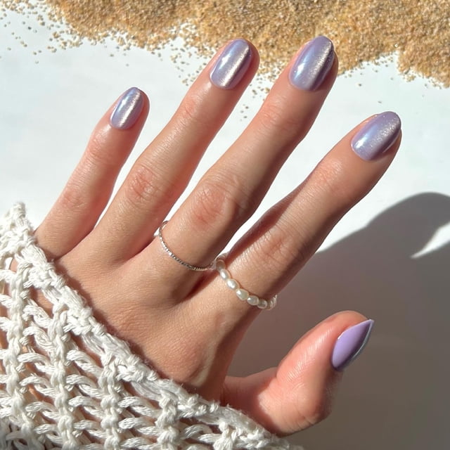 press on nails glamnetic sirenita con forma de cola de sirena corta ovalada, color morado — mano frontal con uñas postizas cortas ovaladas en púrpura iridiscente y acabado brillante tipo terciopelo; reflejos perlados a la luz y forma que evoca cola de sirena, fondo claro con suéter tejido. Ideal para manicure nocturna.