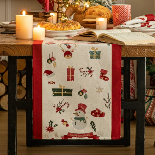 Christmas Table Runner, Winter Christmas Decorations Table Runners 72 Inches Long by Mas Decor displayed on a rustic wooden table. The runner features festive holiday designs such as snowmen, gift boxes, candy canes, and ornaments on a white background with red borders. Ideal for Thanksgiving and fall tableware, it complements indoor and outdoor holiday gatherings. Surrounding the table are candles, baked goods, and Christmas-themed decor, creating a warm and inviting holiday atmosphere.