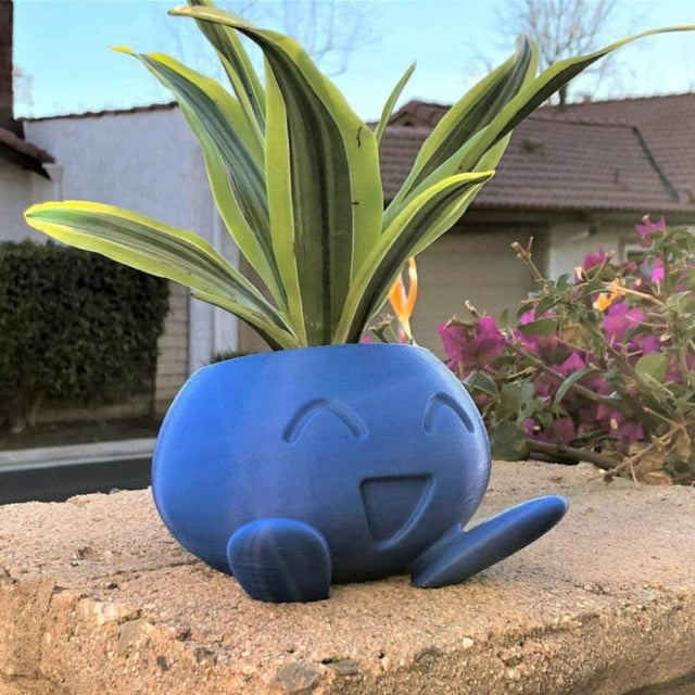 oddish pokemon azul pequeña maceta suculenta maceta brillar electrónica maceta redondeada Oddish azul de frente, ojos y boca sonriente en relieve, patas delanteras modeladas y acabado mate; suculenta variegada plantada en el interior; sobre repisa exterior con flores y tejados desenfocados. Ideal para plantar suculentas o plantas aéreas y como regalo ligero y fácil de limpiar.