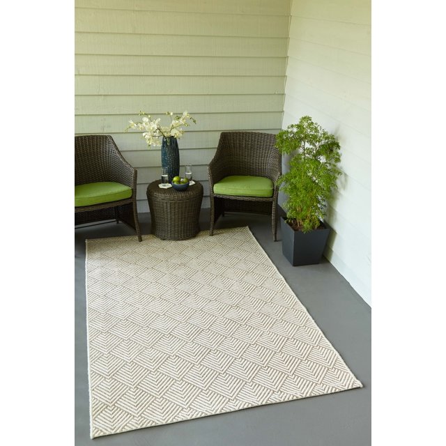 ECARPET Laura contemporary area rug featuring textured geometric diamond patterns in beige, displayed on an outdoor patio floor between two brown wicker chairs with green cushions and accompanied by a small round wicker table and potted greenery.