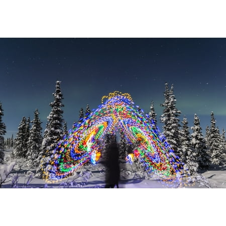The Aurora Borealis dances over the top of a vibrant spiral light painting the blurry figure of a man in the middle of the light painting moonlight casting shadows on snow covered spruce trees on a