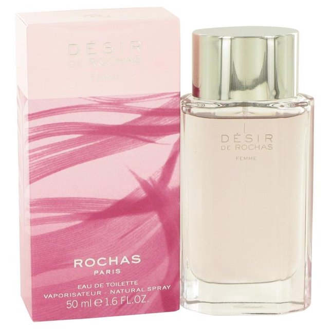 Desir De Rochas by Rochas, Eau De Toilette Spray 1.7 oz for women. The bottle features a sleek, transparent design with a silver cap, showcasing the light pink fragrance inside. The packaging includes a pink box with floral accents and branding details. This floral fruity fragrance has top notes of litchi, black currant, mandarin orange, and wild strawberry; middle notes of casablanca lily, magnolia, freesia, peach, and rose; and base notes of sandalwood, amber, patchouli, and dark chocolate.