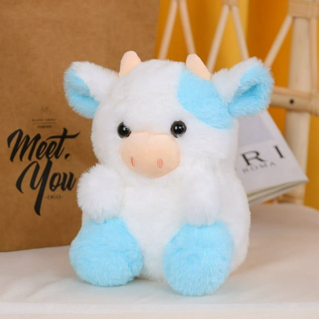 lindo juguete de peluche de vaca, regalo de juguete de peluche para niñas y niños Vista frontal centrada de vaca de peluche multicolor; cuerpo blanco aterciopelado con manchas, orejas y patas azul claro, hocico salmón y ojos negros, silueta sentada sobre superficie interior con bolsa y libro al fondo; ideal para abrazos, juego y acompañar al niño en la siesta.