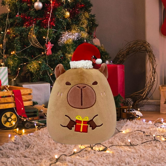 peluche navideño, muñeco de peluche, adorno para sofá, recuerdo de fiesta navideña, muñeco de peluche para abrazar, para adultos y niños. regalos de forma de capibara — vista frontal centrada de peluche capibara de 25 cm con felpa marrón claro; rostro y patitas bordadas que sostienen un regalo rojo con lazo amarillo; lleva gorro navideño rojo con borde blanco; colocado sobre alfombra frente a árbol de Navidad y luces, ideal como adorno para sala de estar.