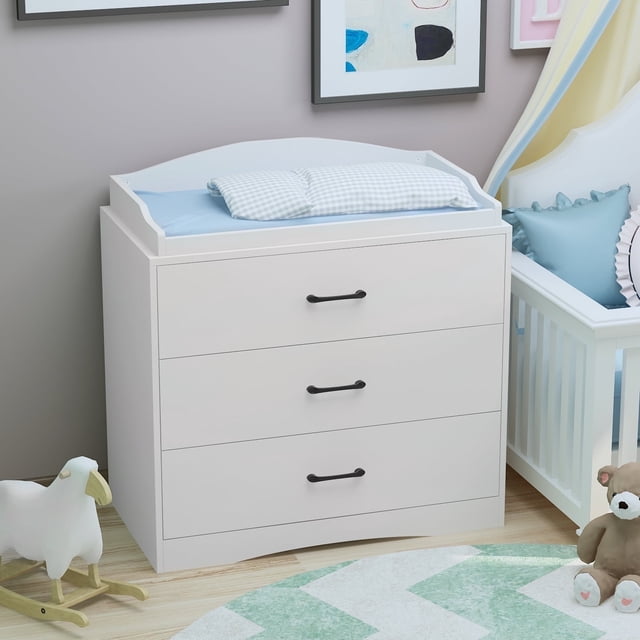 OxyLife Table à Langer Commode, Commode de Chambre de Bébé à 3 Tiroirs avec Dessus à Langer, Blanc. une Pièce de Mobilier de Chambre de Bébé Polyvalente avec Trois Tiroirs Spacieux avec Poignées Noires, un Dessus à Langer Amovible et une construction en Bois Robuste. Idéale pour Organiser les Essentiels pour Bébé. Exposée dans une Chambre de Bébé avec Décor pastel, un Lit de Bébé et des Jouets.