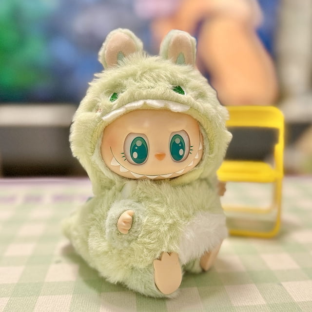 ropa para muñecas con colgante de labubu, generación 2, serie labbu sit party macaron — Conjunto de ropa multicolor para muñeca, vista frontal centrada de disfraz de peluche con capucha con orejas y parche blanco en el vientre; textura tipo felpa, diseñado para muñecas de vinilo kawaii. Fondo interior desenfocado con silla amarilla. También apto como accesorio decorativo para exhibir muñecas pequeñas.