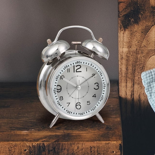 reloj despertador analógico silencioso macarena esfera luminosa para habitación de niños mesita de noche, vista frontal centrada de reloj metálico color argén con doble campana y asa superior, esfera luminosa circular con números arábigos grandes, agujas y segundero de barrido silencioso, patas de apoyo, colocado sobre mesita de noche de madera en dormitorio infantil; también suele usarse como decoración de estantería.