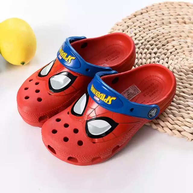 sandalias miniso con dibujos animados, zapatillas rojas para niños y niñas, zapatos casuales de playa, transpirables, de gelatina, para jardín, zapatos de playa de eva ahuecados — Par 3/4 frontal de sandalias infantiles rojas tipo clog con dibujo de ojos estilo superhéroe en la puntera y correa trasera azul con texto amarillo; cuerpo perforado en EVA y plantilla moldeada texturizada, sobre fondo claro; aptas para juego al aire libre y uso en playa, tallas 3–12+.