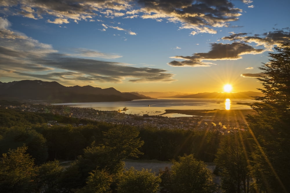 The sun rises over the port of Ushuaia in Argentina as a ship enters the calm bay trees in the foreground and low hills on the horizon while the sky is dotted with clouds and turns from a golden yello
