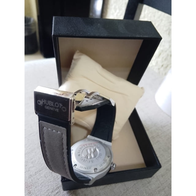 reloj para caballero con esfera gris y cuarzo — reloj Hublot con esfera gris, ventana de fecha a las 3 y bisel plateado; correa de piel con cierre desplegable marcado 'Hublot Geneve', sobre cojín en estuche abierto, vista 3/4 trasera.