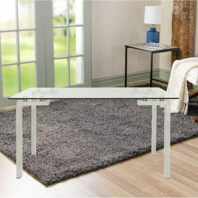 Emma Dining Table in Chrome with a 60-inch rectangular clear tempered glass top and stainless steel tube legs, set on a gray shag rug over light wood flooring near glass-paneled doors and a black metal side table with a blue lamp, suitable for dining or use as a modern desk.