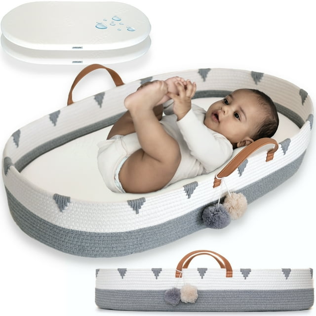 Vesta Baby Cotton Rope Portable Moses Baby Changing Basket Set in gray and white with boho design. The basket features vegan leather handles, pom-pom accents, and a soft foam pad with a waterproof bamboo jacquard cover. A baby is lying comfortably inside the basket, which is suitable for diaper changes or playtime. Includes an extra removable pad cover for convenience.