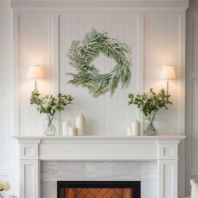 decoraciones navideñas corona navideña para puerta decoración de flores artificiales guirnalda verde multicolor con efecto de escarcha, vista frontal centrada sobre repisa de chimenea en sala minimalista, ramas y follaje texturizado; diseñada para colgar en puerta o pared y a menudo usada también como adorno para mantel durante la temporada.