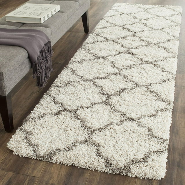 Safavieh Hudson Jaye white shag area rug with gray geometric pattern placed on wooden floor beside a modern gray bench with book and purple throw.