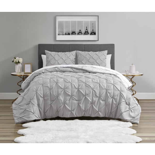 3 Piece Reversible Grey Pinktuck/Paris Revsersible Comforter Set on a bed with a grey upholstered headboard. The light grey pintuck side faces up, showing textured folds, while the reverse side with a white Paris-themed print is partially visible. Two matching pintuck pillow shams rest against the headboard. The room features wood flooring, a white shag rug at the bed's foot, and gold-trimmed round side tables with decorative accents. A framed black and white Eiffel Tower photo hangs on the wall behind.