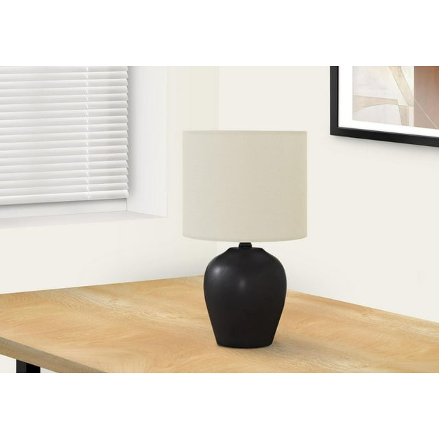 Monarch Specialties Lighting 17"h table lamp with a smooth black ceramic round jar base and ivory fabric drum shade on a light wood corner table near a window with white blinds and framed abstract art, suitable for bedroom or office use.