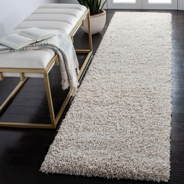 Light beige plush shag runner rug on dark hardwood floor beside white cushioned bench with gold frame, styled with open book and striped throw.