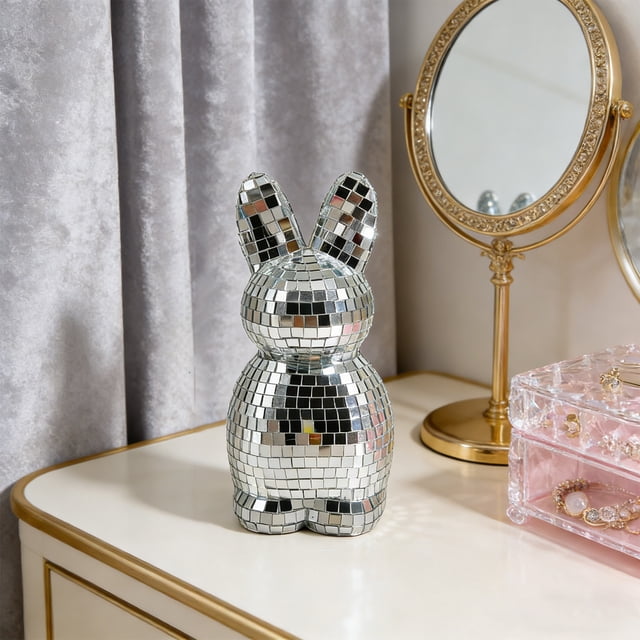 adorno de conejo de pascua con forma de conejo, espejo, estatua de conejo hecha a mano Blanco — figura decorativa de conejo recubierta de pequeños azulejos de espejo tipo mosaico, vista frontal centrada sobre una cómoda blanca; silueta redondeada con orejas largas y superficie reflectante facetada, situada junto a un espejo de tocador dorado y una caja organizadora rosa en un interior doméstico; adecuada también como acento decorativo para mesa de noche o escritorio.