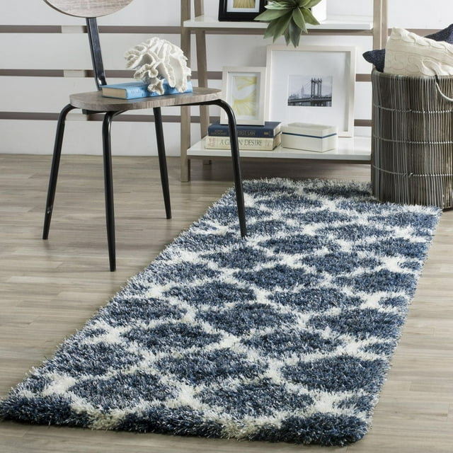 Safavieh Montreal Lennart blue and white geometric shag area rug displayed in a modern room setting with wooden floor, black metal chair, decorative coral piece, books, framed photos, plant on shelf.