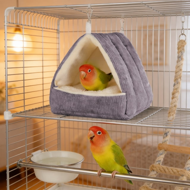 hamaca cálida para loros | cama colgante de algodón grueso y afelpado en gris con interior crema, vista frontal 3/4 en jaula; refugio triangular acolchado con base mullida, costuras acanaladas y dos anillas metálicas superiores para colgar; un loro dentro y otro posado en el borde junto a comedero y perchero. Espacio acogedor para descanso invernal de loros medianos, también adecuado como cama transportable para cortos periodos fuera de la jaula.