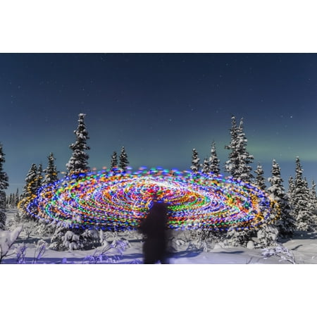 The Aurora Borealis dances over the top of a vibrant spiral light painting the blurry figure of a man in the middle of the light painting moonlight casting shadows on snow covered spruce trees on a