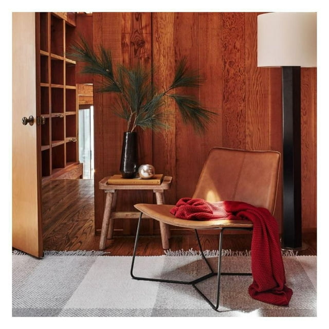 Plata Décor Slope Chair in tan PU leather with black metal frame, styled with a red knitted throw in a cozy living room setting featuring wood-paneled walls and rustic decor.