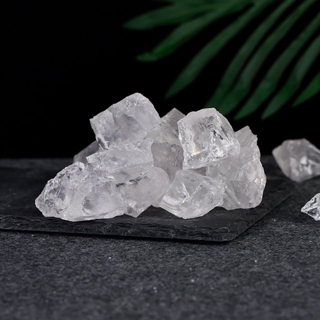 piedra de cristal en bruto, piedra de aromaterapia, piedra perfumada, polvo natural, cristal verde fluorita, amatista, citrino, piedra de cristal — Montón frontal de piezas de cristal en bruto blancas translúcidas sobre una losa negra; toma en 3/4 que muestra bordes angulares, caras facetadas naturales y vetas internas; fondo negro con hoja verde desenfocada, adecuada para aromaterapia, spa o como recuerdo de viaje.