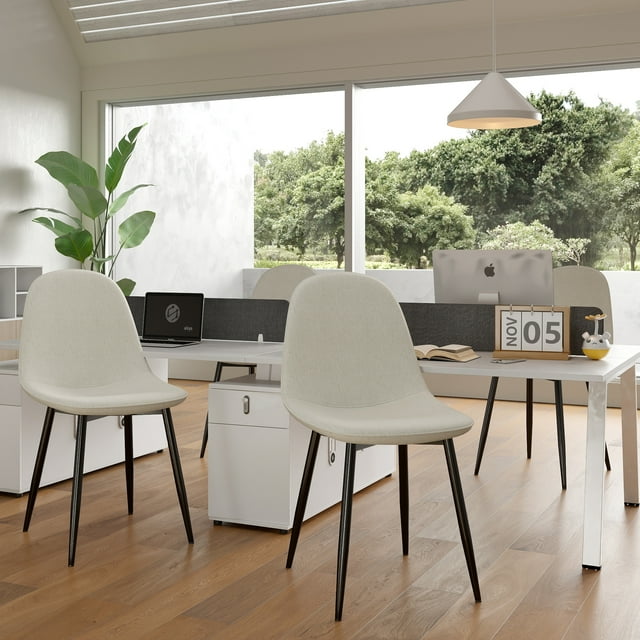 Four beige fabric upholstered armless office side chairs with black metal legs arranged around a white desk in a modern home workspace with large windows and wooden flooring.