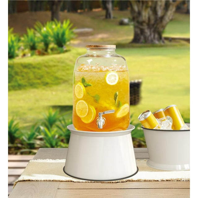 Hometrends White Round Dispenser Stand, Dia 9.56" galvanized steel base supporting a glass beverage dispenser filled with iced lemon and mint drink on a wooden outdoor table with a white runner, next to a white ice bucket with yellow cans, suitable for outdoor entertaining.