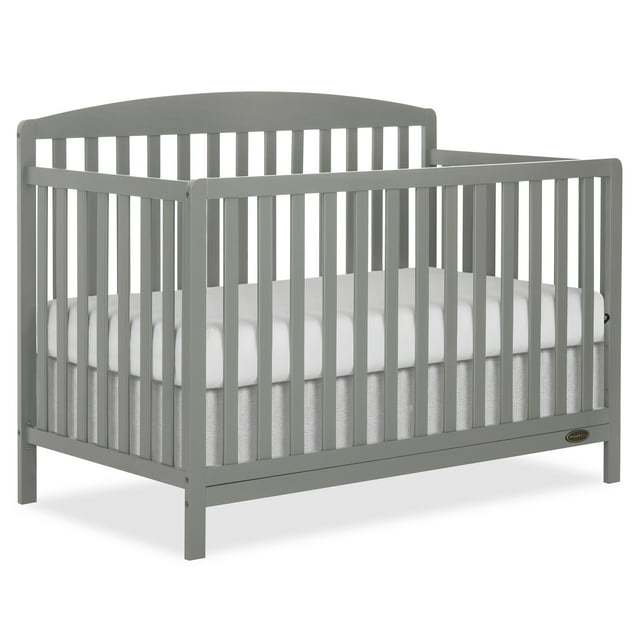Dream On Me Odelle 5 in 1 Convertible Crib in sea grass finish, shown in a 3/4 front view with vertical wooden spindles and an arched headboard, featuring a white mattress and grey crib skirt, made from sustainable New Zealand pinewood with non-toxic water-based paint, includes 3-position adjustable mattress height and converts to toddler and full-size beds for nursery use.