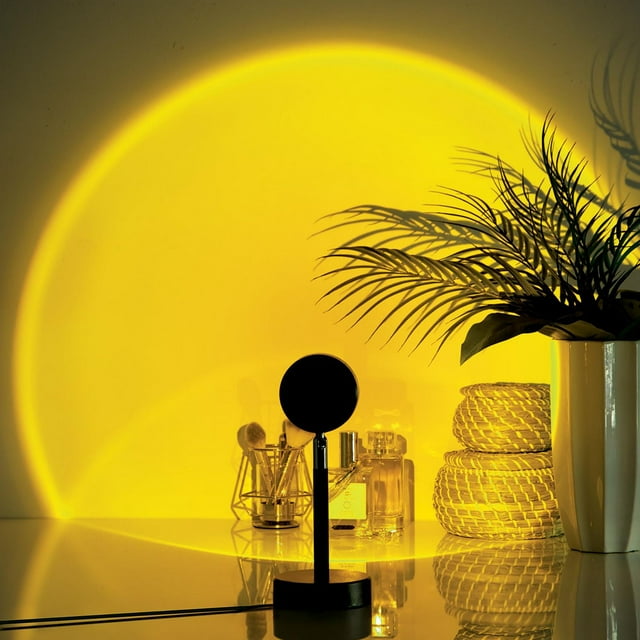 Merkury 4-in-1 sunset lamp LED projector casting warm yellow light on wall with makeup brushes, perfume bottles, woven baskets and potted plant.