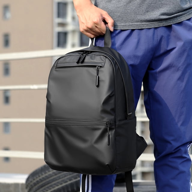 mochila grande para hombre, impermeable, para escuela de negocios, portátil, mochila informal (negro) 3/4 frontal de una mochila negra rectangular de poliéster (42×30×15 cm) con superficie suave y acabado mate, cremallera superior, bolsillo frontal con cremallera y tiradores de goma, asa superior y tirantes acolchados; apta para laptop de 14 pulgadas; fondo urbano desenfocado sugiere uso diario y oficina; también usada como bolso de hombro.