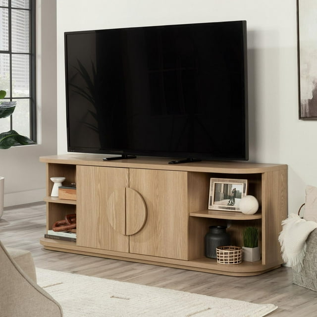 Sauder Select TV Stand in Harmony Ash Finish holding a large flat-screen TV, with two central cabinet doors and open shelving for decor or media storage.
