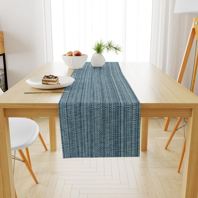 Blue woven cotton Fabstyles casual classic table runner on light wood dining table with white chairs, dessert plate, and decorative vases in bright room.