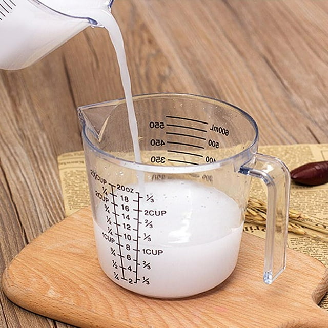 taza medidora de plástico ligero con asa y boquilla medida de de leche de agua de harina para la preparación de alimentos 600 ml 3/4 frontal con leche vertiéndose; jarra medidora transparente de 600 ml con pico y asa recta, marcas impresas en ml y tazas en negro, plástico claro sobre tabla de madera en encimera. Ideal para medir líquidos y secos al hornear; frecuentemente usada para preparar mezclas pequeñas.