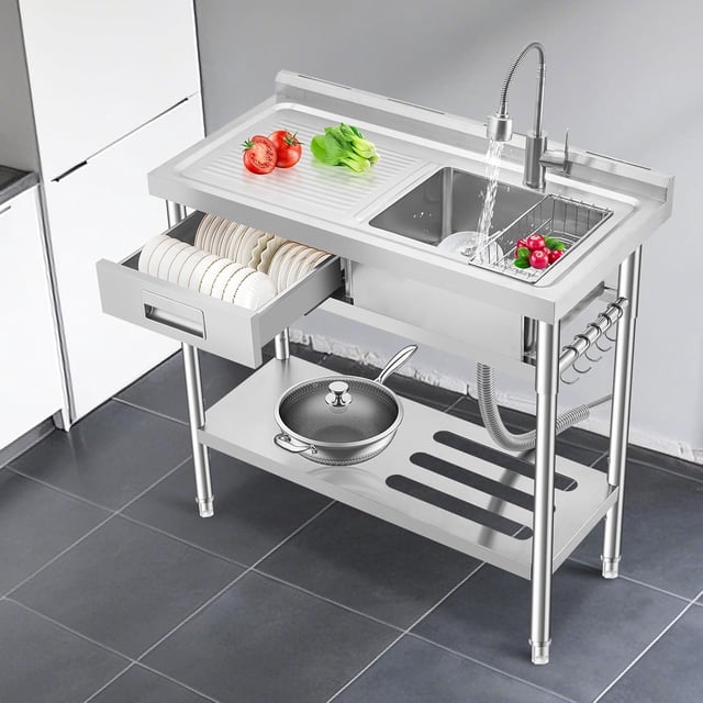 historyli fregadero independiente de acero inoxidable con banco de trabajo, cajón izquierdo y grifo de cocina comercial Vista 3/4 frontal del fregadero Historyli en acero inoxidable con encimera integrada y escurridor a la izquierda, cuenca rectangular con grifo de cuello flexible y agua en funcionamiento; cajón izquierdo en forma de L abierto con platos alineados, estante inferior para ollas y tuberías visibles, ganchos laterales para utensilios y patas metálicas ajustables sobre piso de baldosas. Suele usarse también como estación de preparación en cocinas pequeñas.