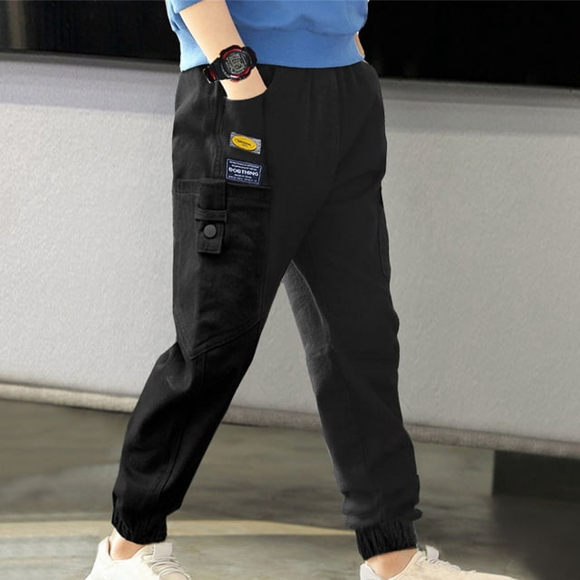 YUTK solid color spring boys pants in black, designed for middle-aged children. These casual workwear pants feature an elastic waistband for easy wear, a side pocket with a button closure, and a decorative patch. Made from soft, breathable, and durable fabric, suitable for indoor and outdoor activities. Shown in a lifestyle setting with a child wearing a blue top and white sneakers.