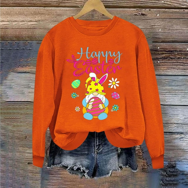 Jsaierl Chandails de Pâques Joyeuses pour Femmes avec un Chandail orange vibrant à Enfiler avec une Impression Graphique de Lapin, Texte « Joyeuses Pâques » et des Motifs Colorés sur le Thème de Pâques comme des Oeufs et des Fleurs. le Chandail à Col Rond, Manches Longues et Coupe Décontractée, Style avec des shorts en denim sur un Fond en Bois Rustique.