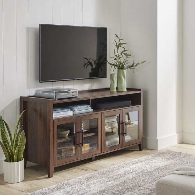 Better Homes & Gardens Reading Refined Farmhouse TV Stand with dark wood finish and four glass-paneled doors with black metal handles. It fits TVs up to 65 inches and features an open console with wire management holes holding a turntable, books, and a soundbar. Positioned against a white paneled wall with a mounted flat-screen TV, decorated with green ceramic vases and plants, on a light wood floor with a patterned rug. Also suitable for organizing media accessories and decor.