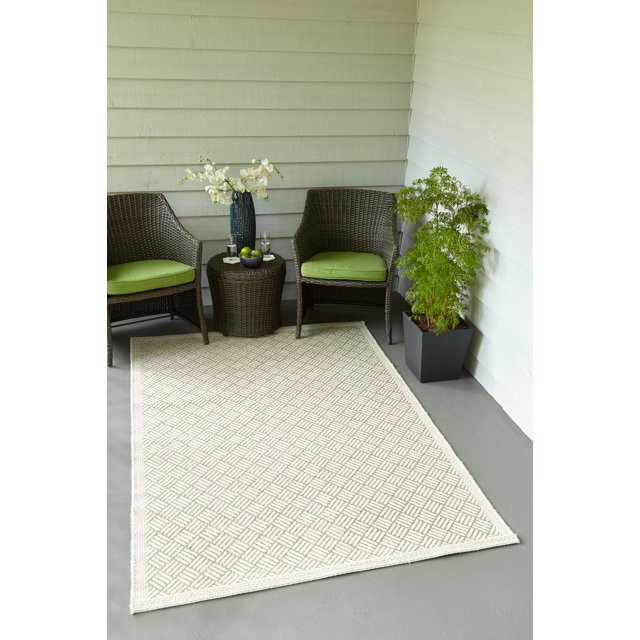 ECARPET Eve contemporary beige geometric patterned area rug on gray floor between two brown wicker chairs with green cushions and round side table holding glasses, fruit bowl, and white flower vase in an outdoor seating space.